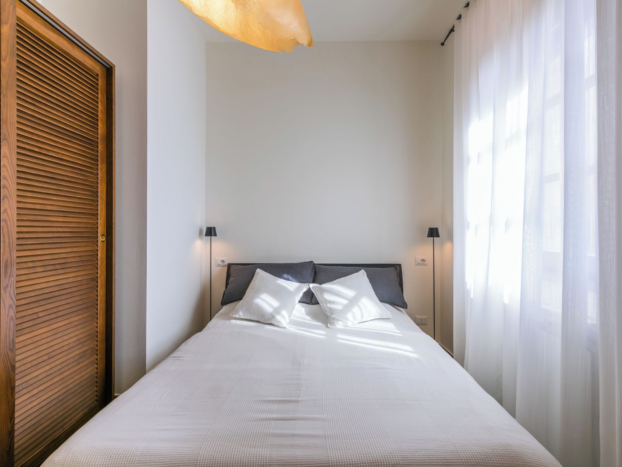 A neatly made bed with a headboard, two bedside lamps, and a window with sheer curtains.