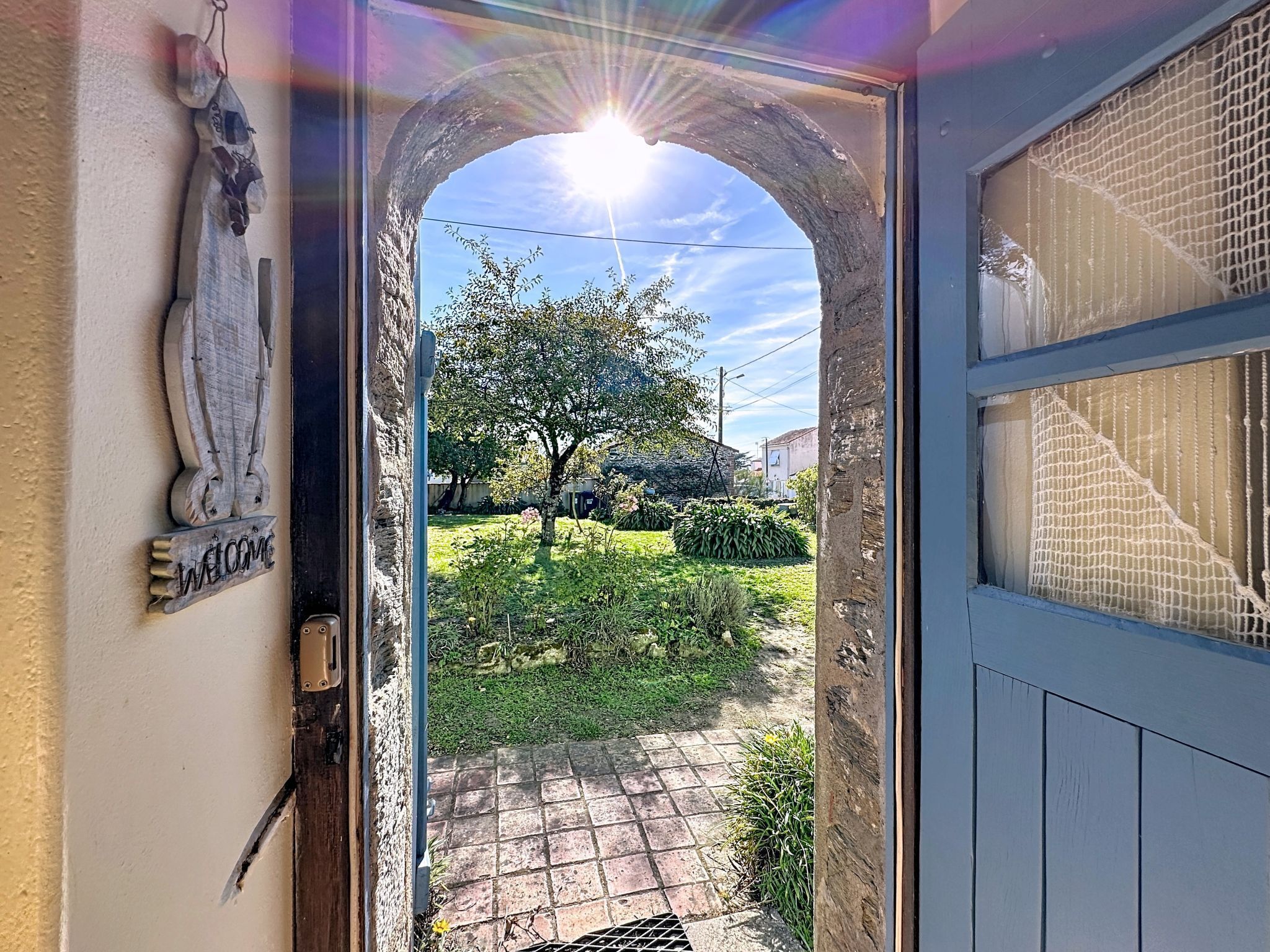 Un arco de entrada a una propiedad, con un letrero de bienvenida, vistas de un jardín y una puerta azul con ventana.