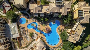 Vue aérienne d’un complexe hôtelier doté d’une grande piscine centrale en forme de guitare, entourée de plusieurs bâtiments avec balcons et d’un espace paysager agrémenté de palmiers.
