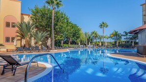 Une piscine entourée de chaises longues et de parasols, bordée de palmiers et d’un bâtiment doté d’un toit en tuiles.