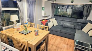 A living room with a wooden dining table, grey sofa, and a TV. - Kingfisher Caravan Park - Holiday Accommodation 22193 (Skegness)