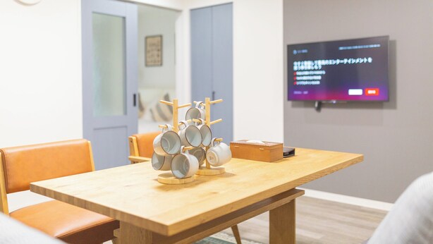 A room with a wooden table, chairs, a clock, and a TV displaying Japanese text. - b&Kishinosato (Osaka)