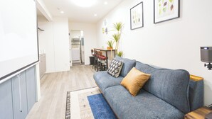 A modern living room with a grey sofa, a blue rug, and a wooden coffee table. There is a kitchen area with a dining table and chairs in the background. - b&Kishinosato (Osaka)
