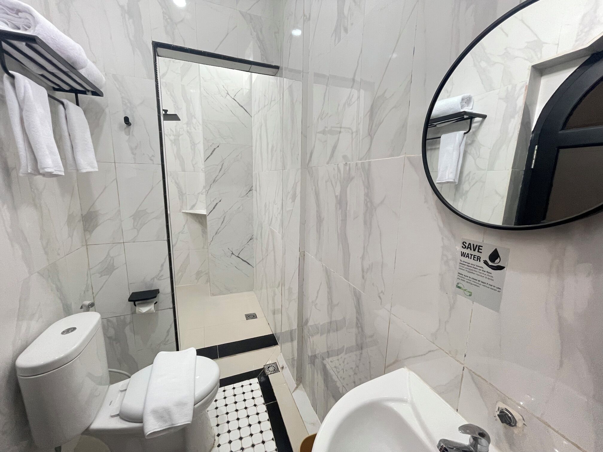 Ein modernes Badezimmer mit Duschkabine aus Glas, Toilette, Waschbecken und Spiegel. Ein Schild mit der Aufschrift 'SAVE WATER' ist an der Wand zu sehen.
