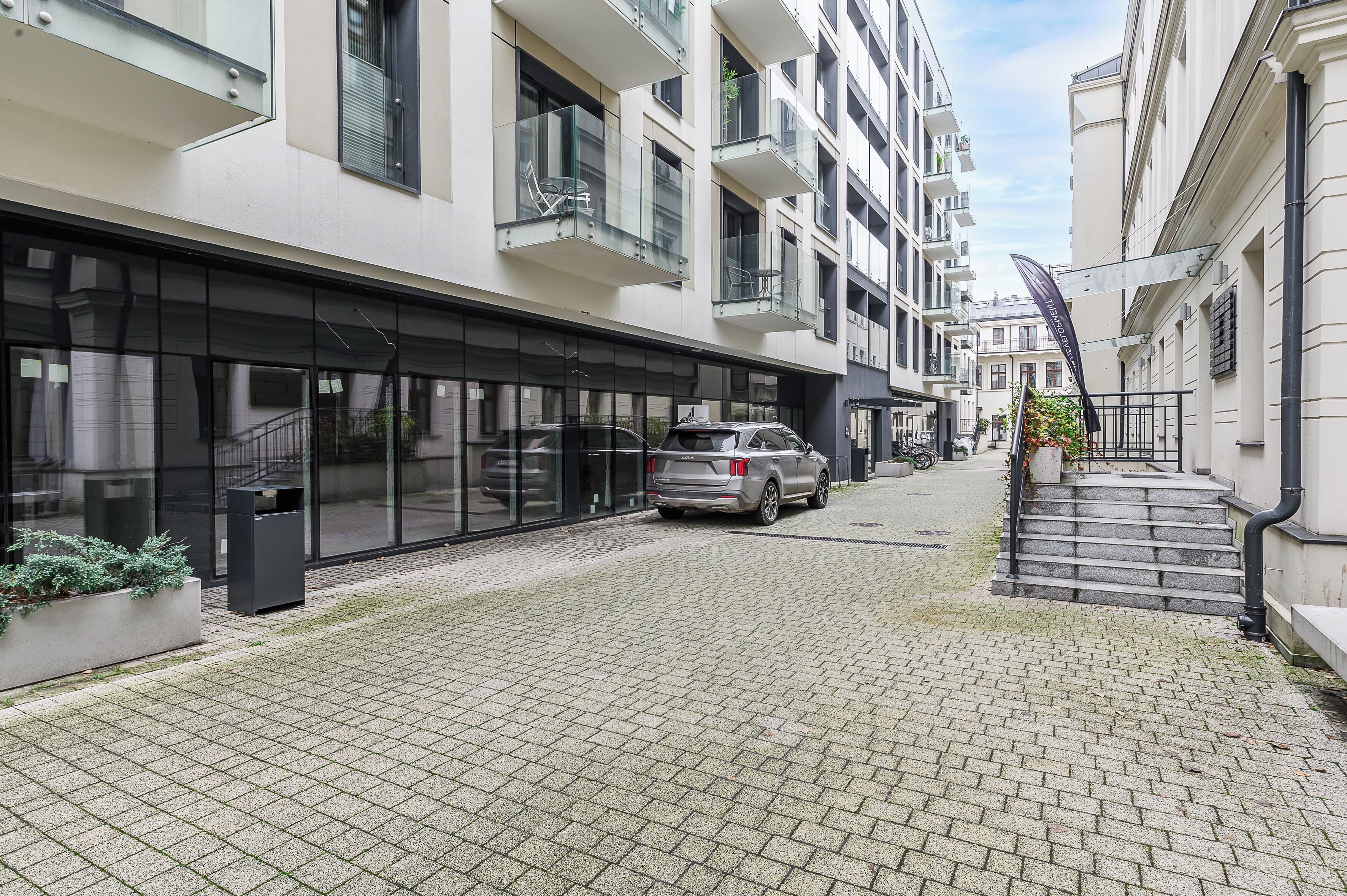 A narrow cobblestone street flanked by modern buildings with balconies, a parked car, and a small garden area.