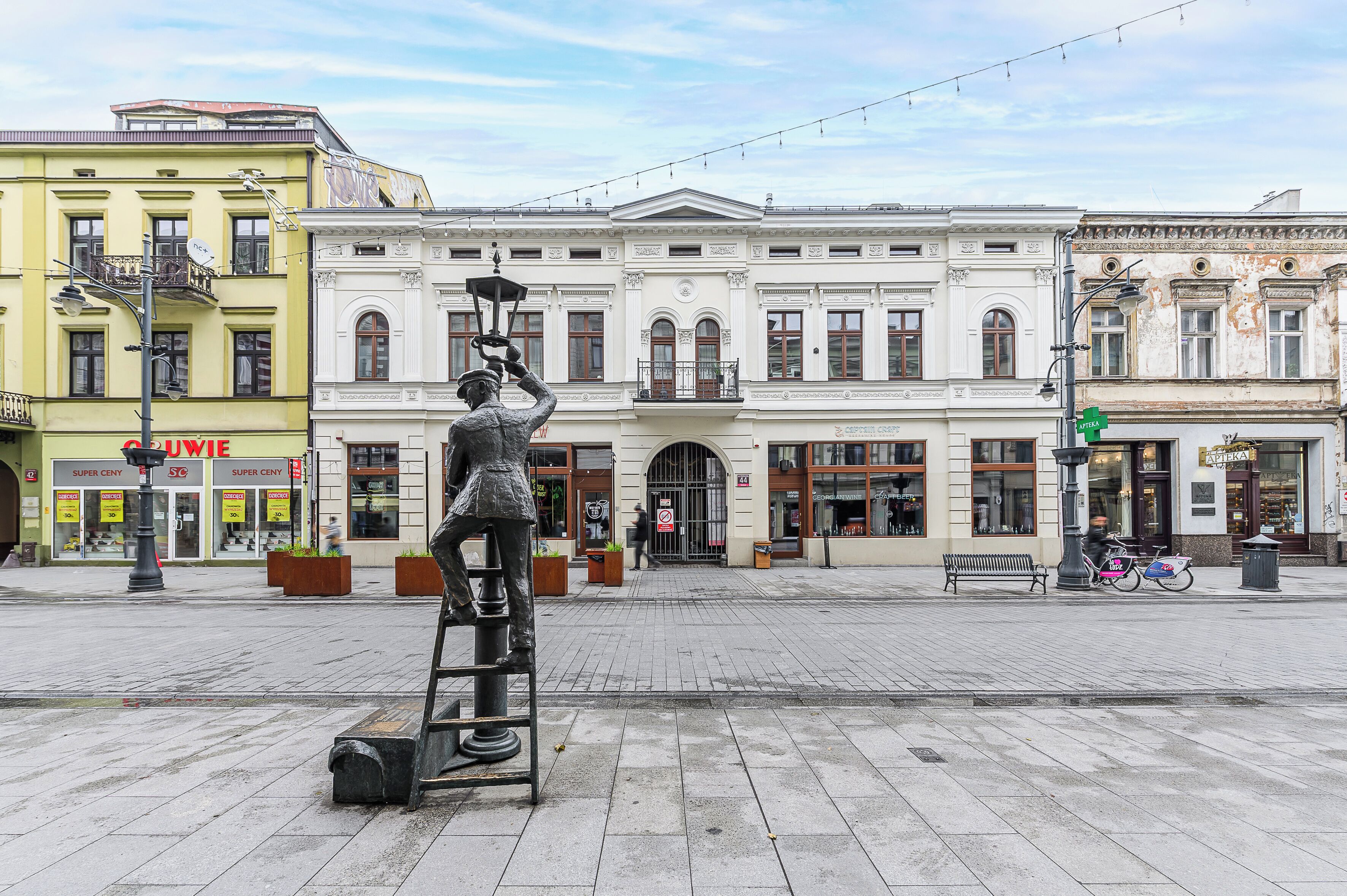 Una plaza pública con una estatua de una persona sosteniendo un farol, rodeada de edificios con diferentes tiendas, incluyendo una tienda de vinos.