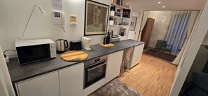 Une cuisine compacte équipée d’un four à micro-ondes, d’un grille-pain, d’une machine à café et d’une planche à découper en bois. On y trouve également un petit four, un évier et une chaise près du comptoir. Un tableau encadré orne le mur, et une porte donne accès à un balcon avec vue.