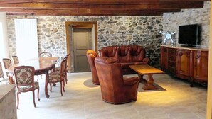 A cozy living room with a stone wall, wooden ceiling beams, a leather sofa, a dining table with chairs, and a television on a wooden cabinet.