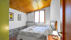 Une chambre avec un plafond en bois, un lit recouvert d’une courtepointe blanche, une table de chevet avec une lampe et une fenêtre avec des rideaux.