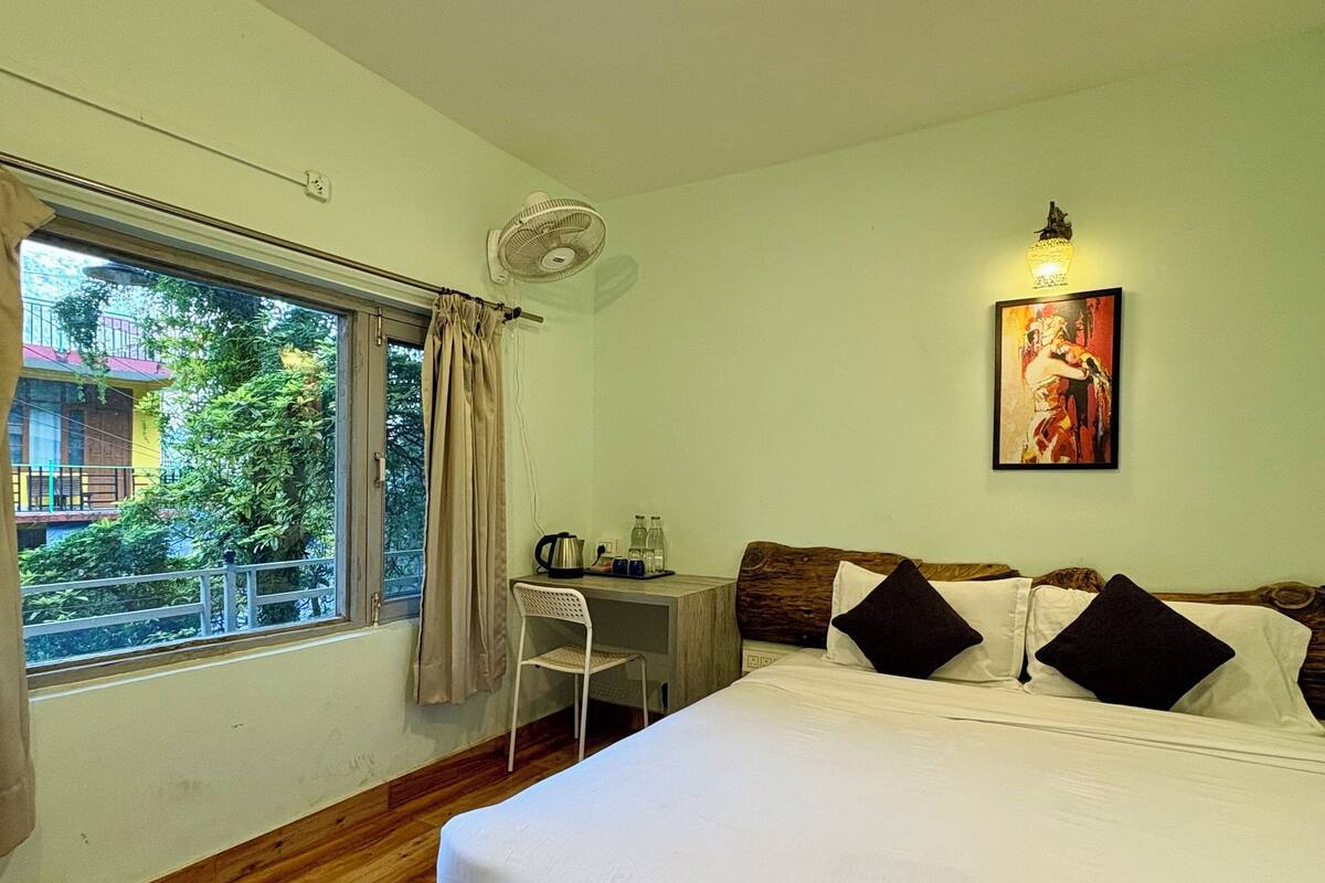 Ein Schlafzimmer mit Holzboden, einem Bett mit weißer Bettwäsche und schwarzen Kissen, einem Schreibtisch mit Stuhl, einem Fenster mit Vorhängen und einem Wandventilator.