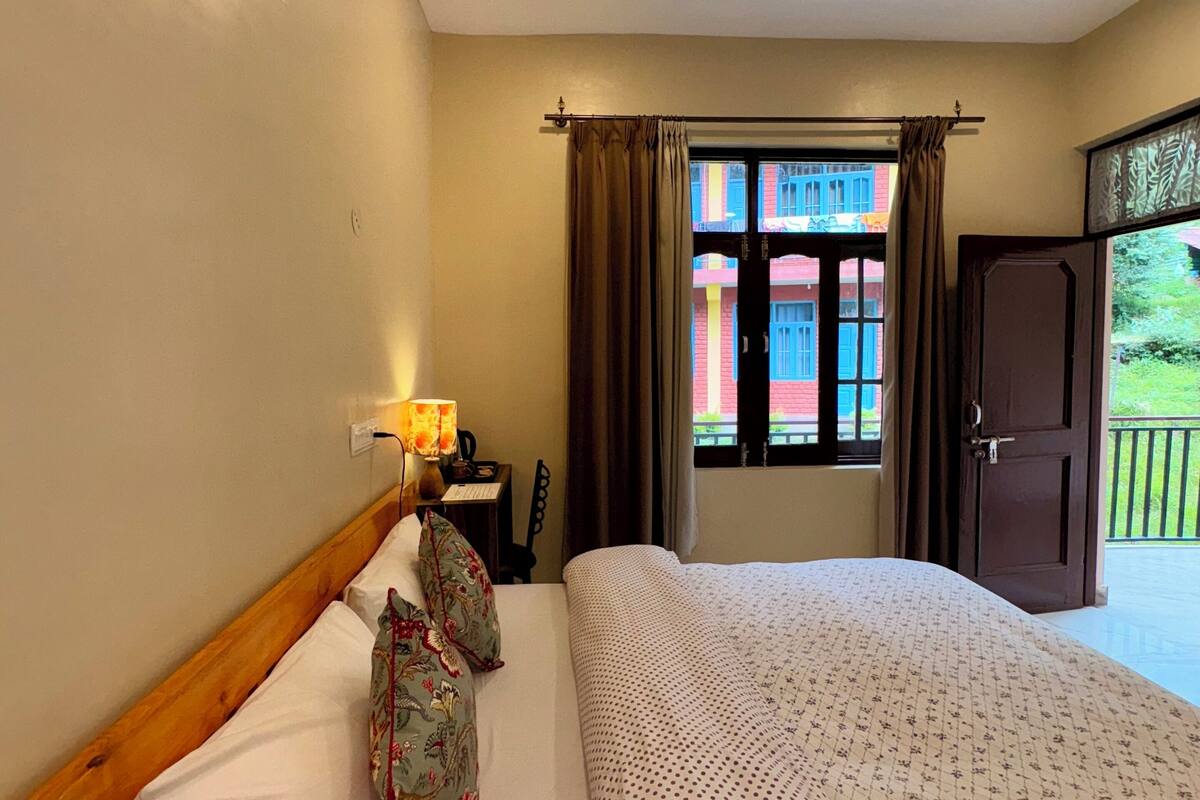 Ein Hotelzimmer mit Bett, Nachttisch, Lampe, einem Fenster mit Vorhängen und einer Tür, die auf einen Balkon führt.