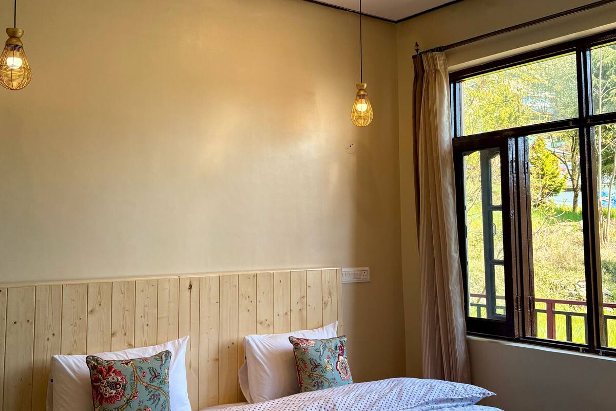 Ein Schlafzimmer mit einem hölzernen Kopfteil, zwei Pendelleuchten, einem großen Fenster mit Vorhängen und einem ordentlich bezogenen Bett mit gemusterten Betttextilien und gerollten Handtüchern.