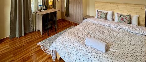 A bedroom with a wooden floor, a bed with floral bedding, a desk with a chair, and a wardrobe.