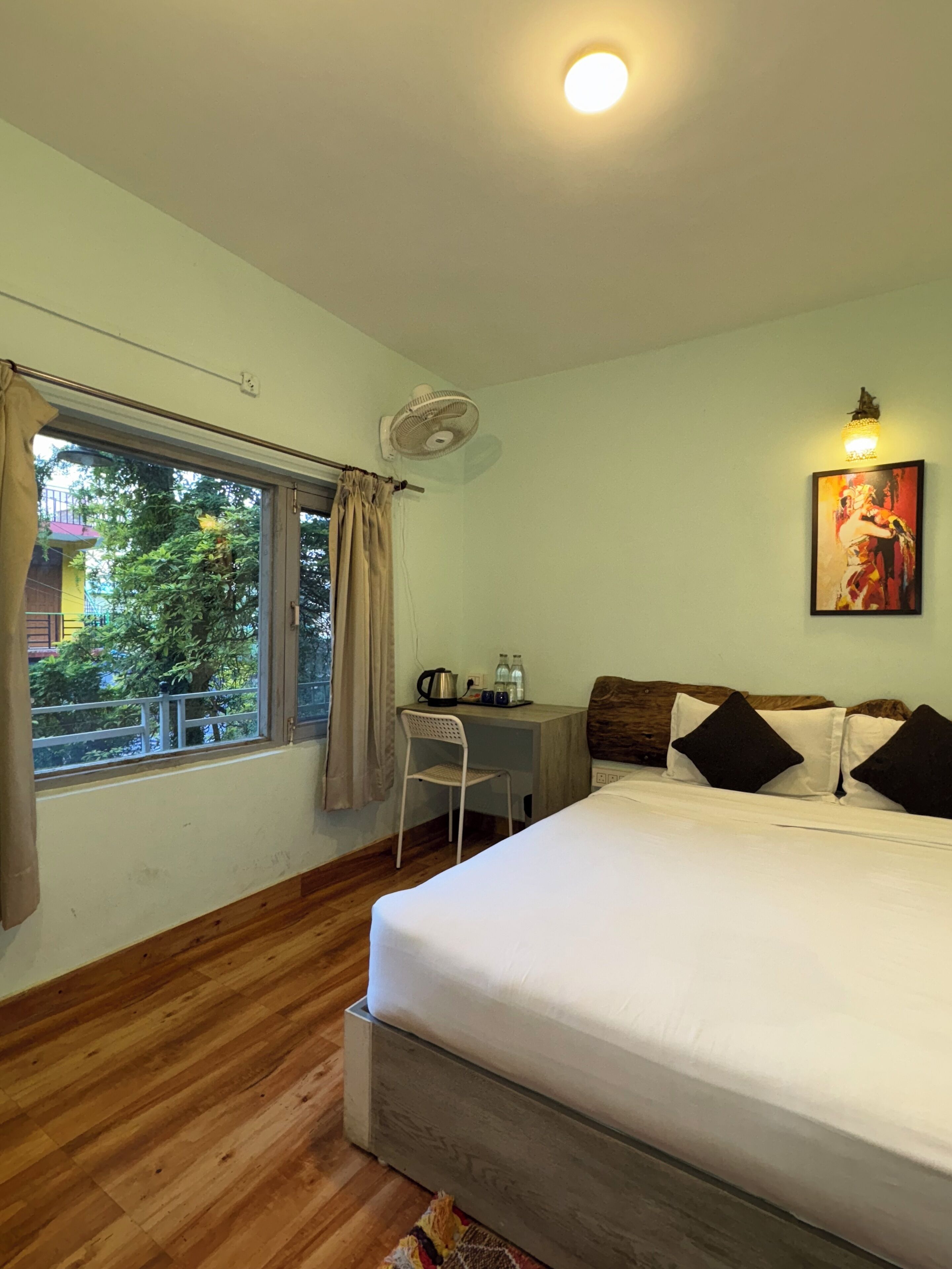 A hotel room with a bed, a desk with a chair, a window with curtains, a ceiling fan, and a painting on the wall.