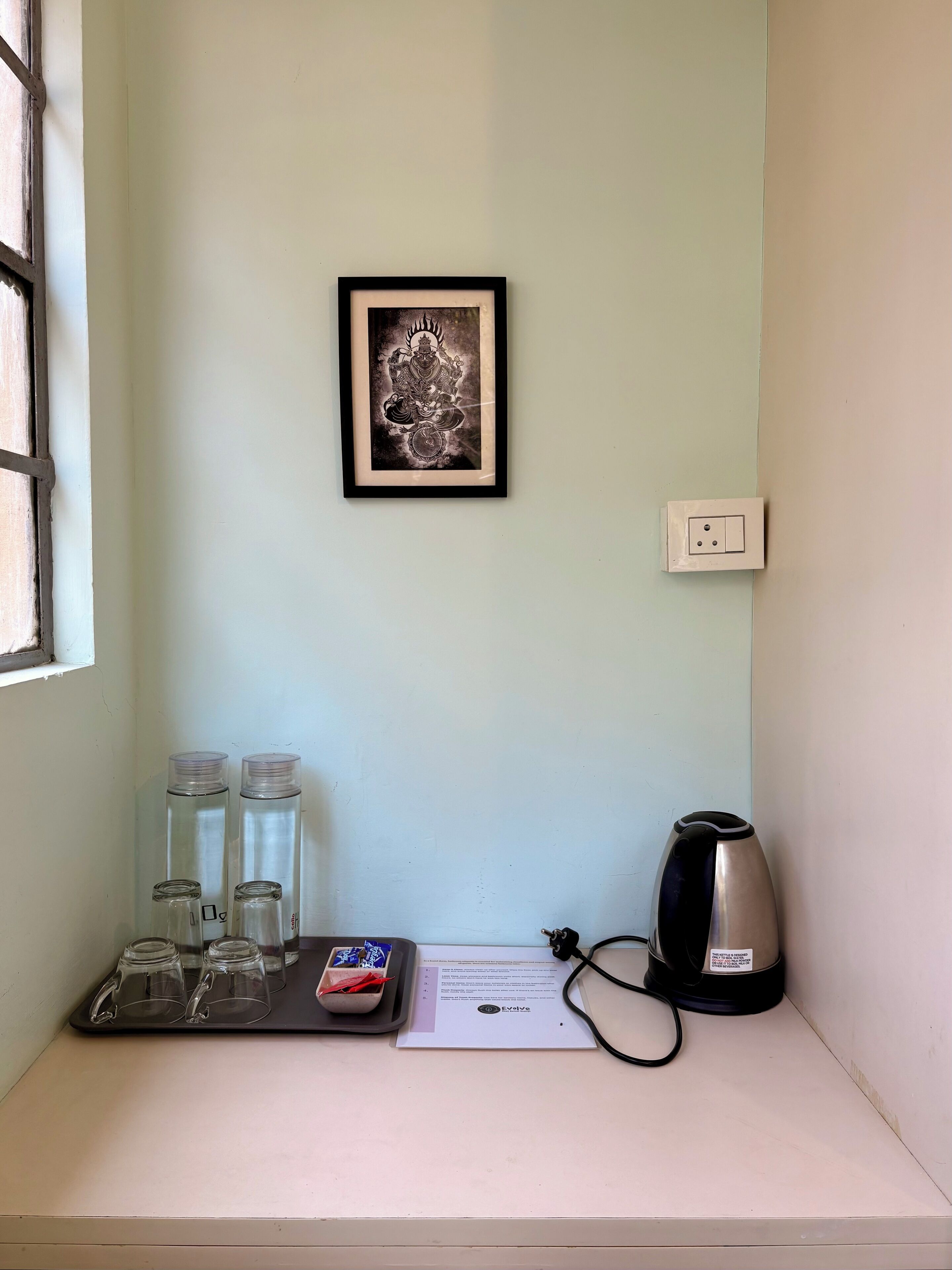 A small room with a framed artwork on the wall, a kettle, and a tray with glasses and a tissue box.