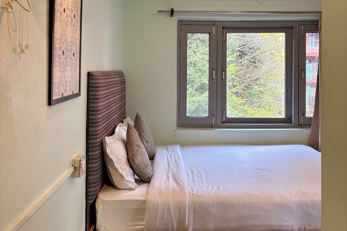Ein Schlafzimmer mit Bett, Nachttisch, Wandventilator, Fenster mit Blick auf Bäume und einem gerahmten Bild an der Wand.