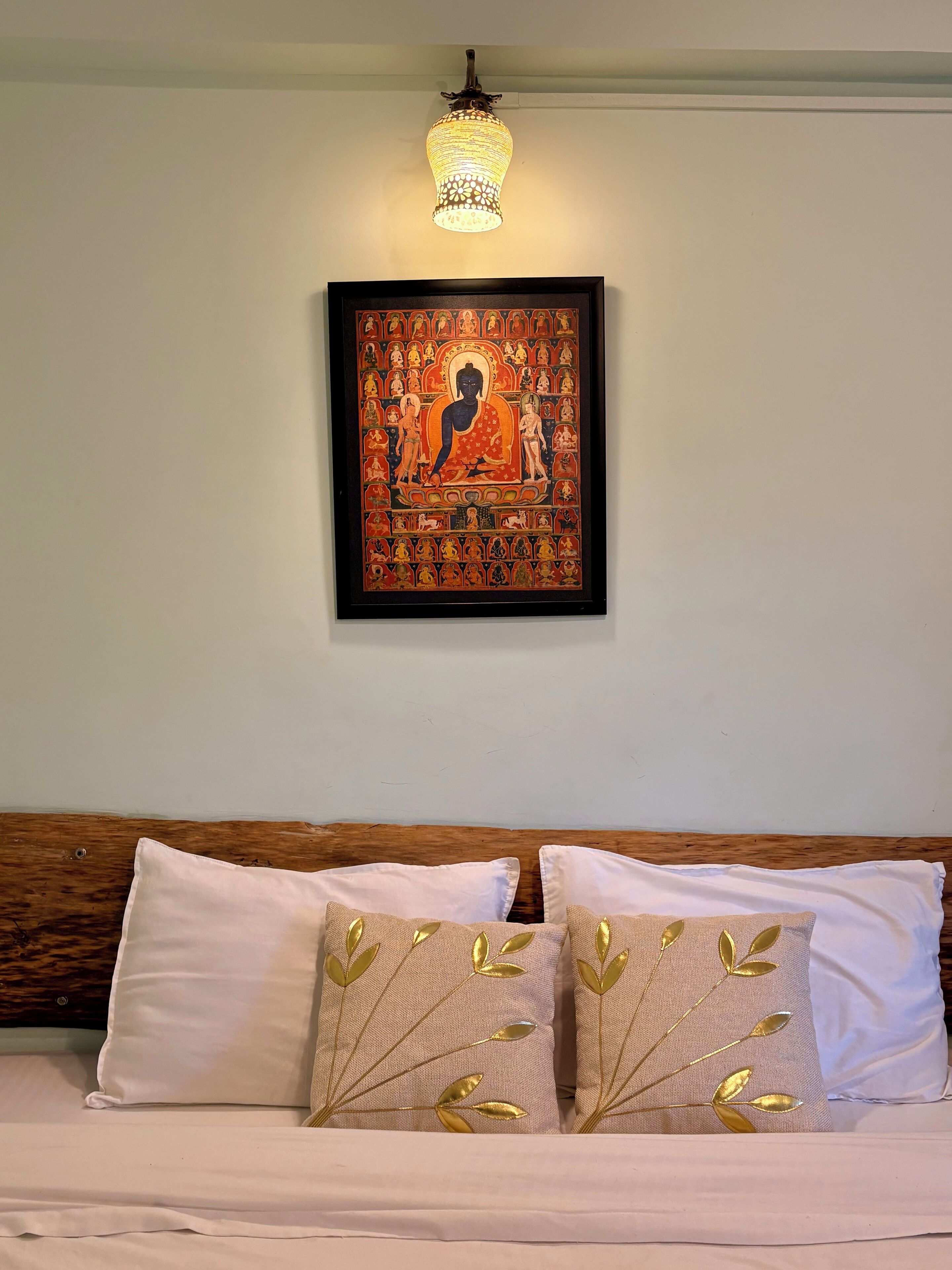 Een bed met witte lakens en twee kussens met bladgoud motieven, een gelijst boeddhabeeld aan de muur en een hanglamp.