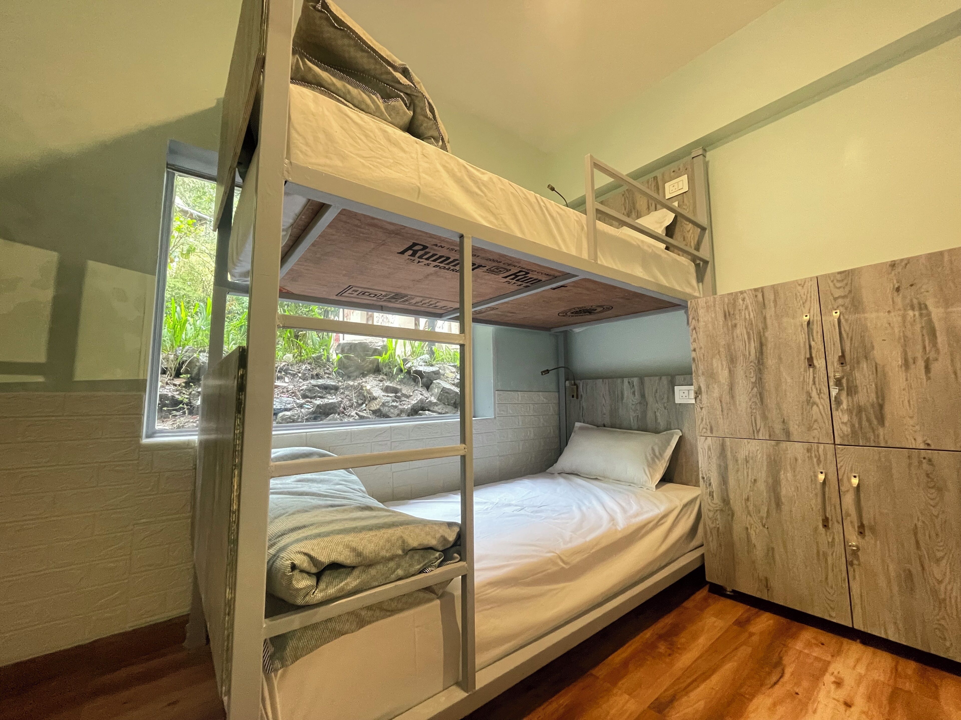 A bunk bed with a window, a wooden wardrobe, and a view of greenery outside.