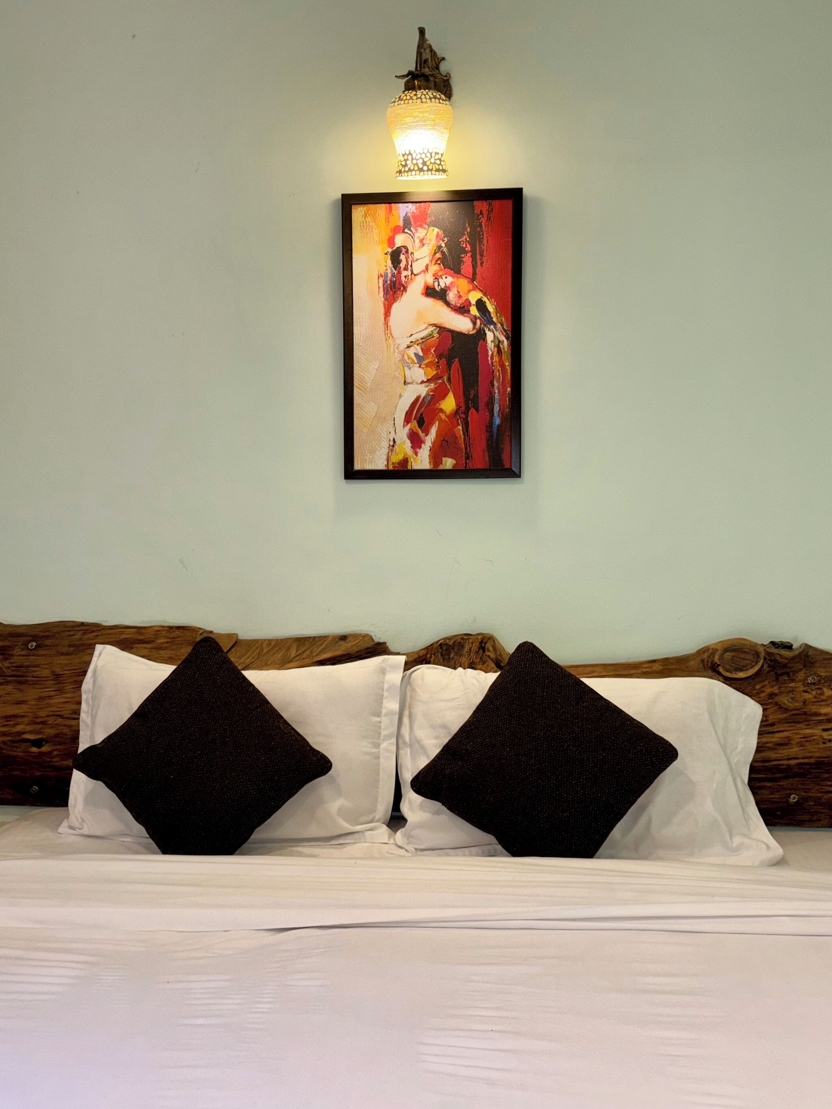A bed with white linens and two black pillows, a wall-mounted light fixture, and a framed painting above the bed.