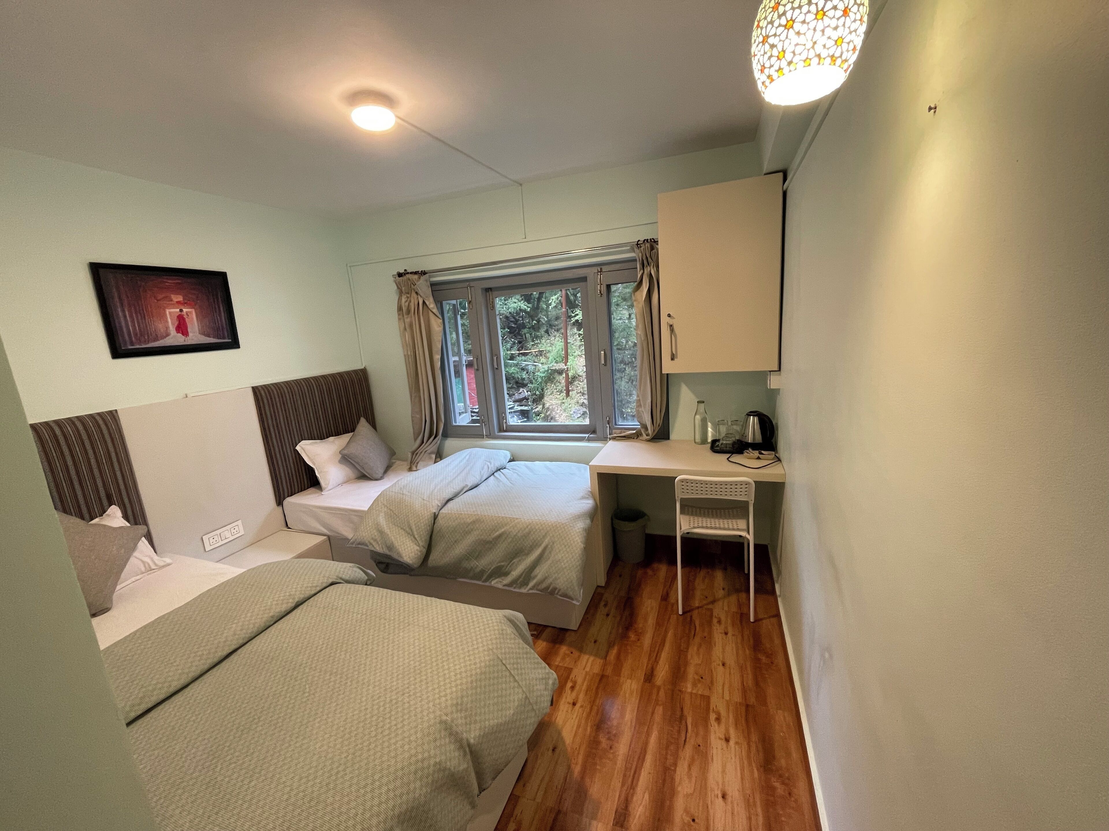 A small, well-lit room with two beds, a desk, and a window with curtains.