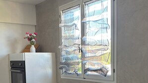 A kitchen with a built-in oven, a refrigerator, a window with patterned curtains, and two bar stools. - La Cachette du Sud  (Vallauris)