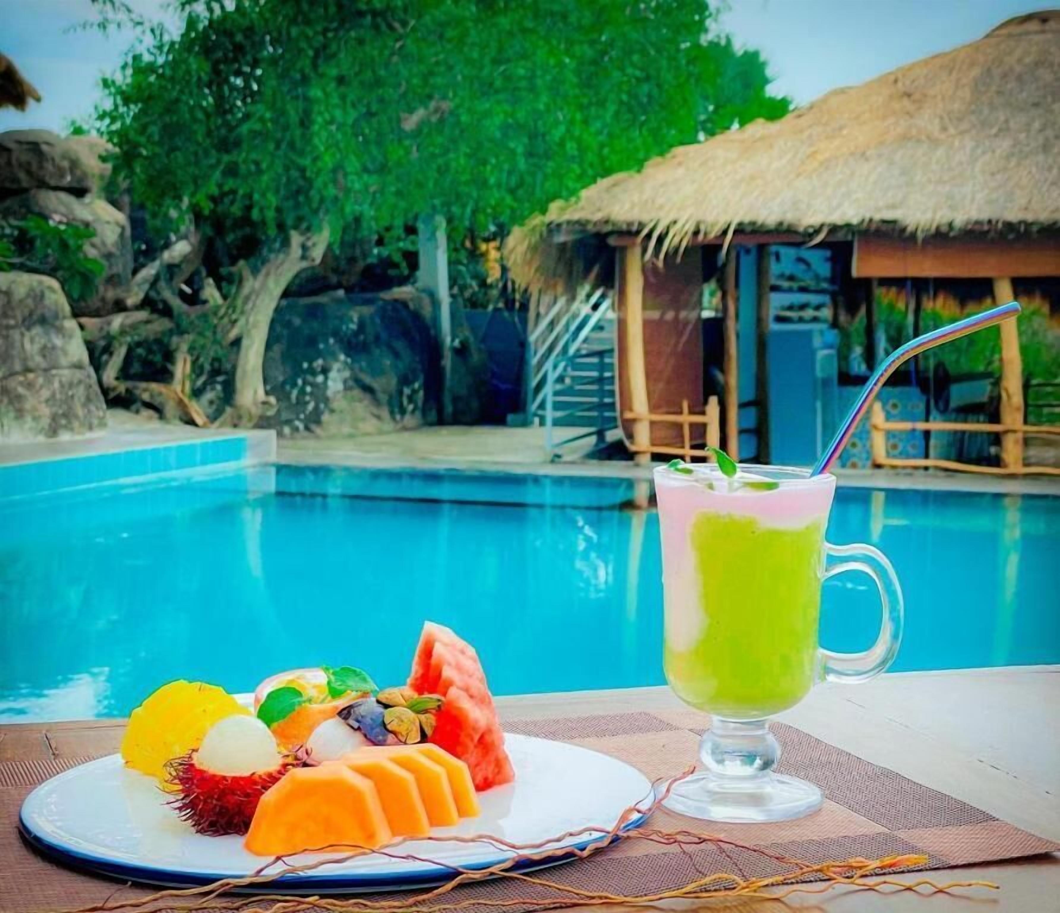 Un cadre agréable au bord de la piscine, avec une assiette de fruits et une boisson verte, un abri à toit de chaume et une piscine à l’eau d’un bleu limpide.