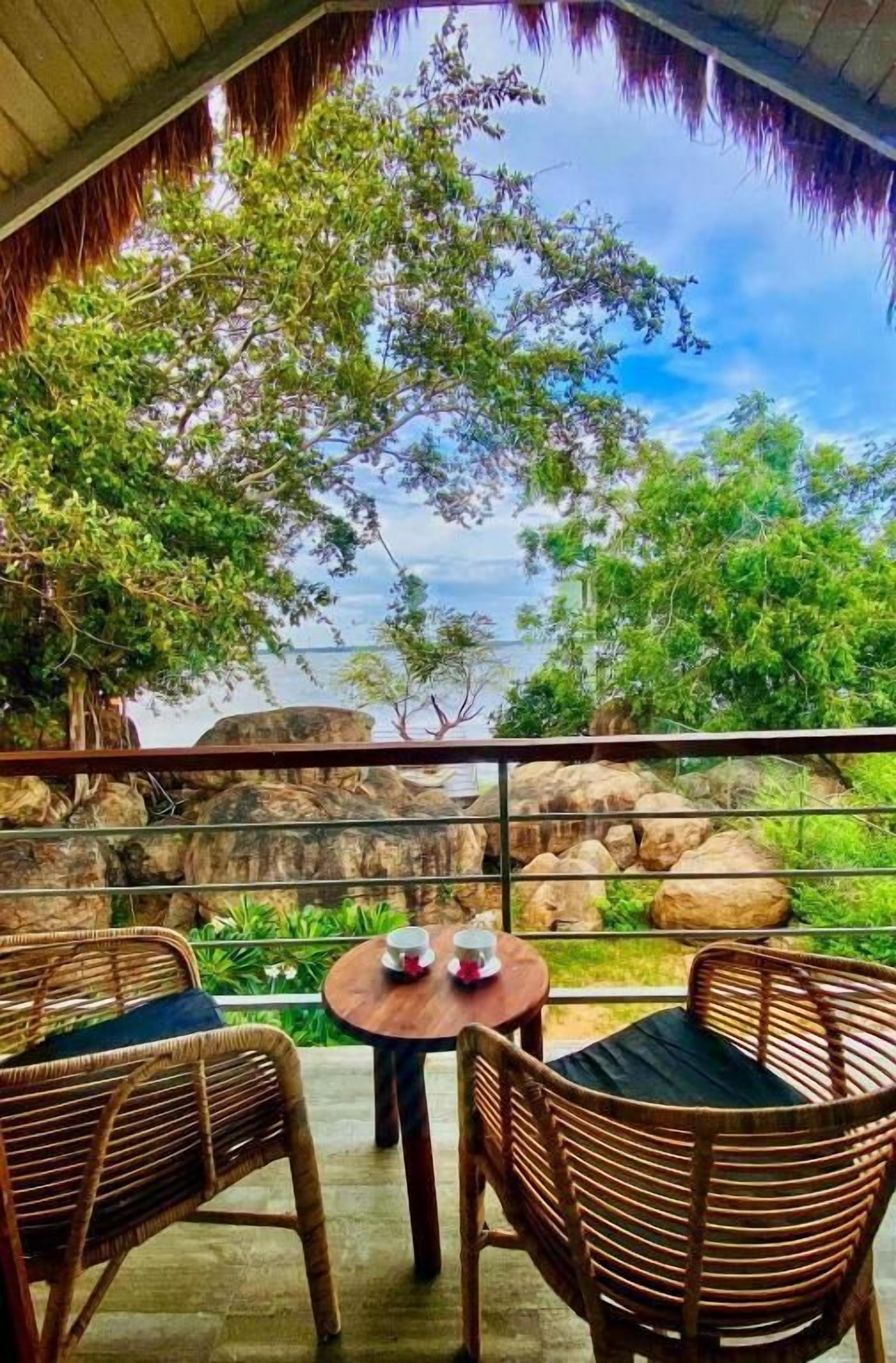 A covered outdoor seating area with wicker chairs and a round table, overlooking a natural landscape with rocks and greenery.