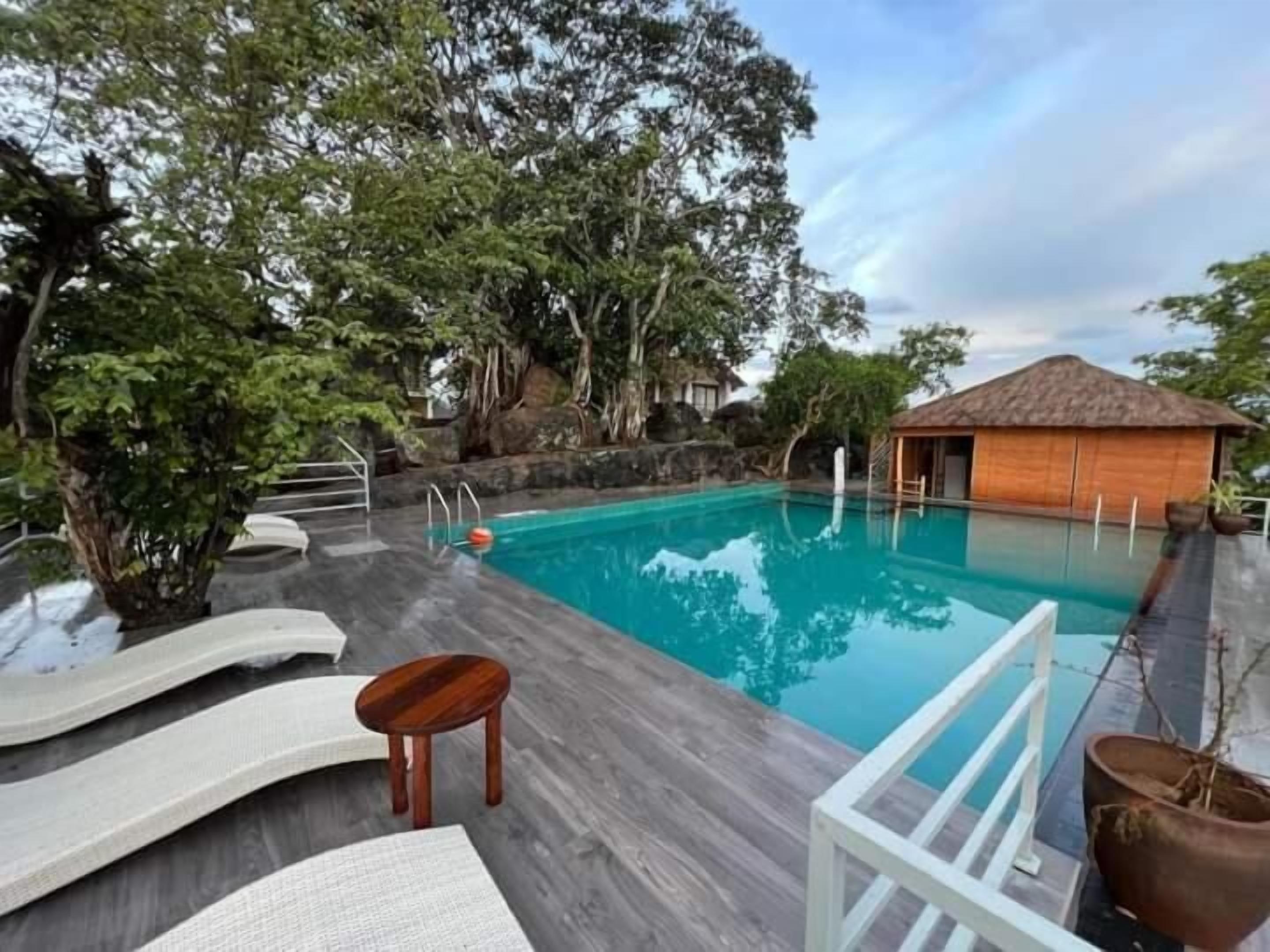 A pool area with lounge chairs, a wooden deck, and a thatched-roof building.