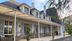 Une maison à deux étages dotée d’un patio couvert, de nombreuses fenêtres avec volets et d’un espace salon extérieur.