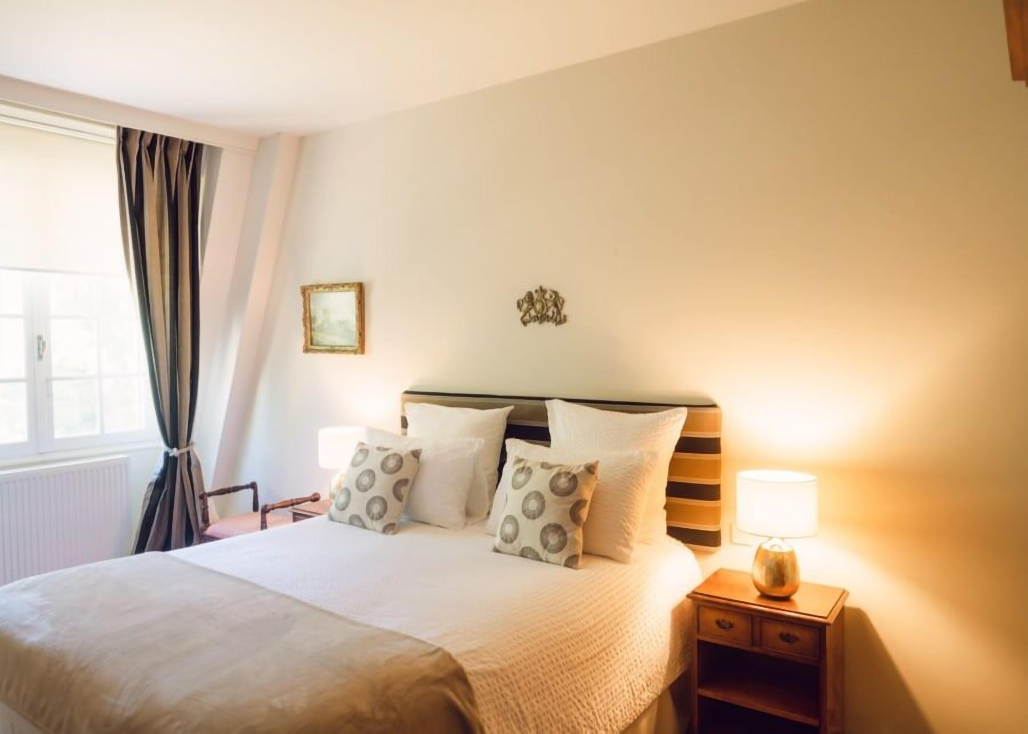 A hotel room with a large bed, a nightstand with a lamp, a window with curtains, and a wall-mounted picture frame.
