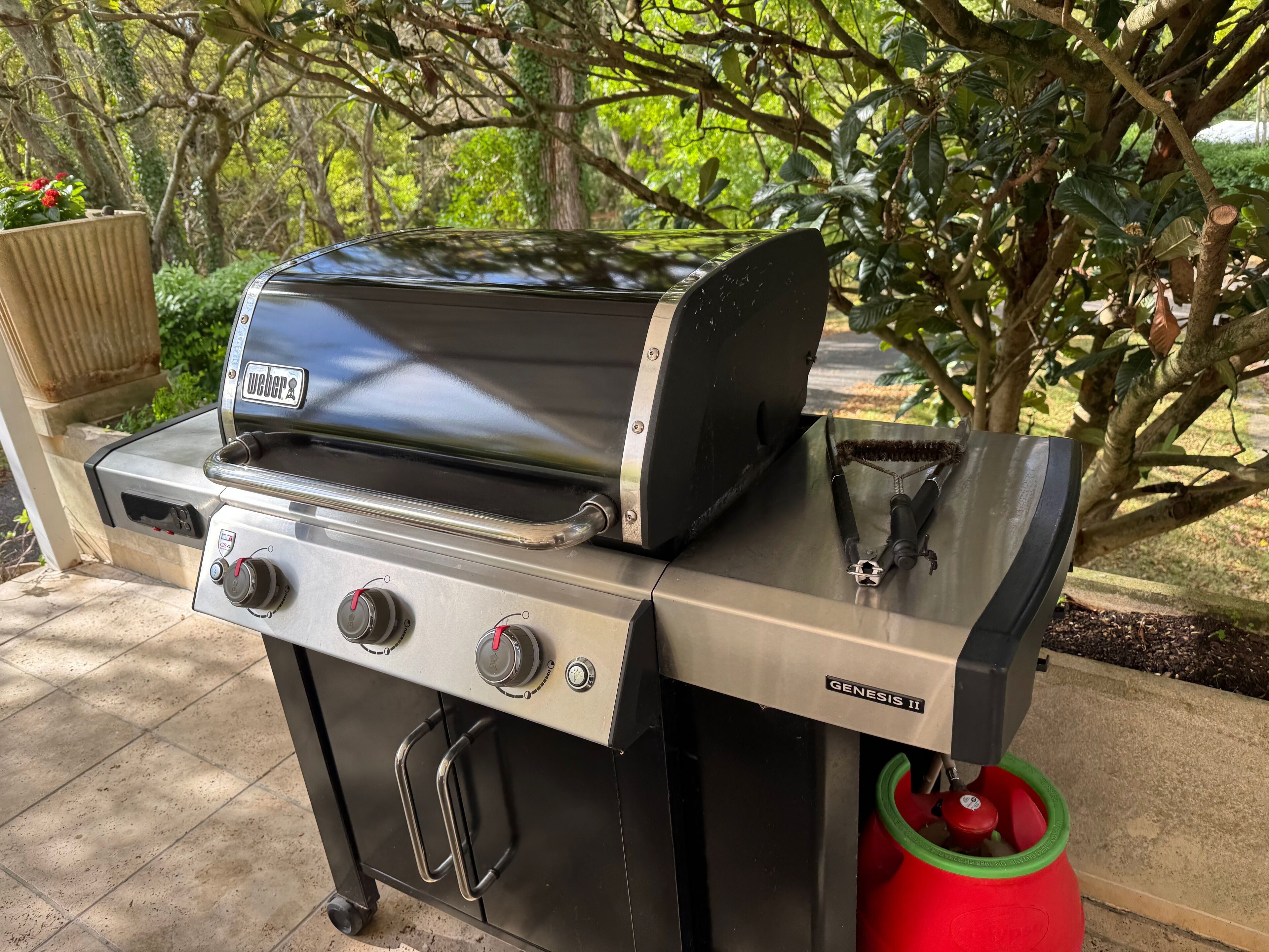 Una parrilla de gas Weber Genesis II con cuatro quemadores, un estante lateral y una bandeja para grasa, ubicada al aire libre con un jardín de fondo.