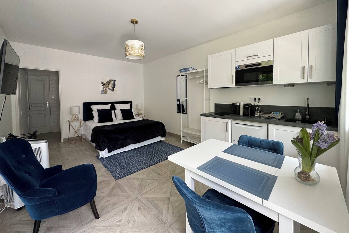 A compact hotel room with a bed, a desk, a chair, a kitchenette, and a TV.