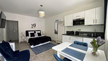 A compact hotel room with a bed, a desk, a chair, a kitchenette, and a TV.