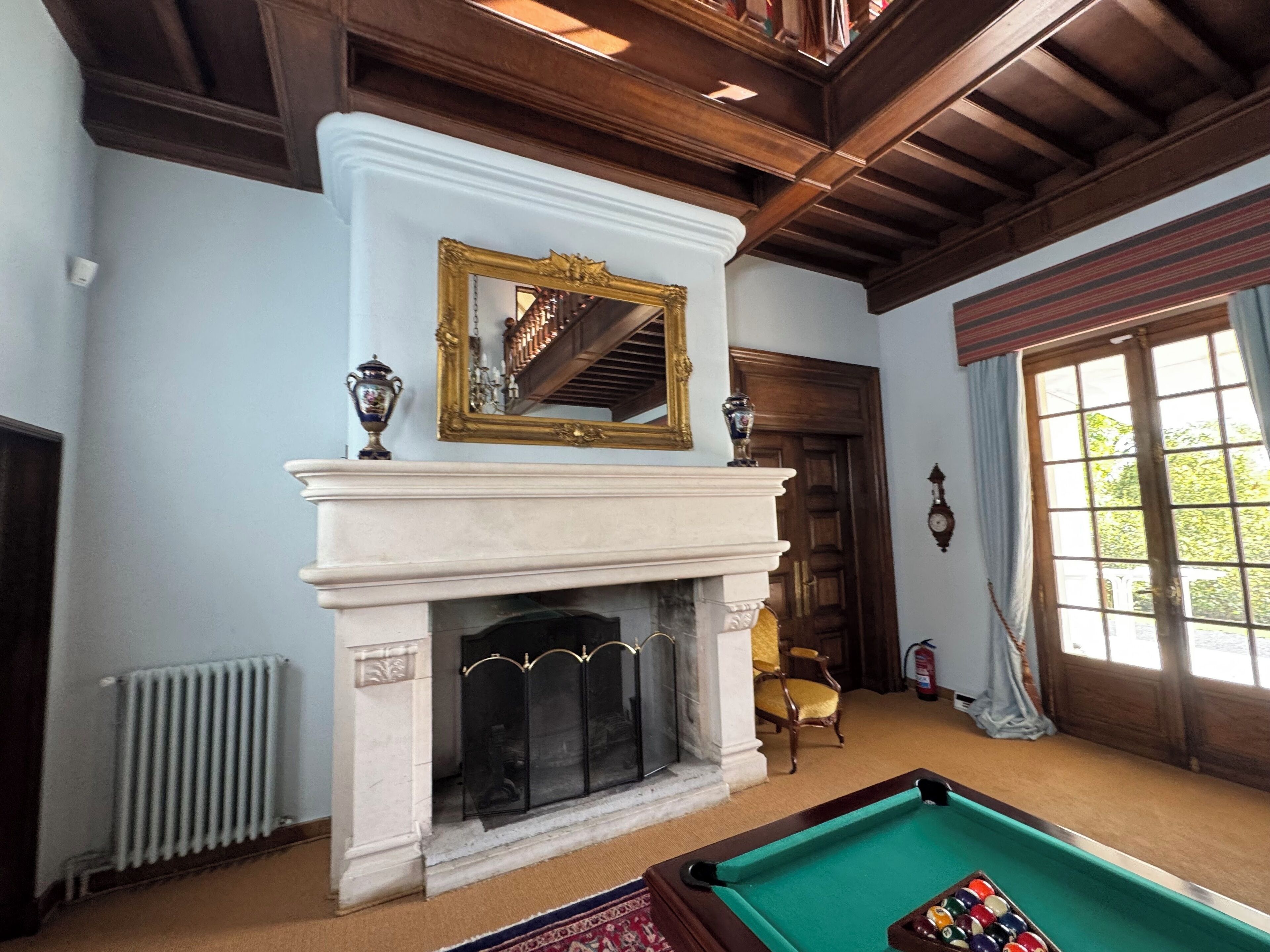 A room with a fireplace, a pool table, a mirror, and a window with curtains.