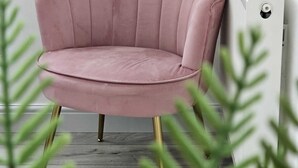 Una butaca rosa tapizada, con respaldo festoneado y patas doradas, colocada frente a un radiador blanco en una habitación con suelo de madera y follaje verde al frente.