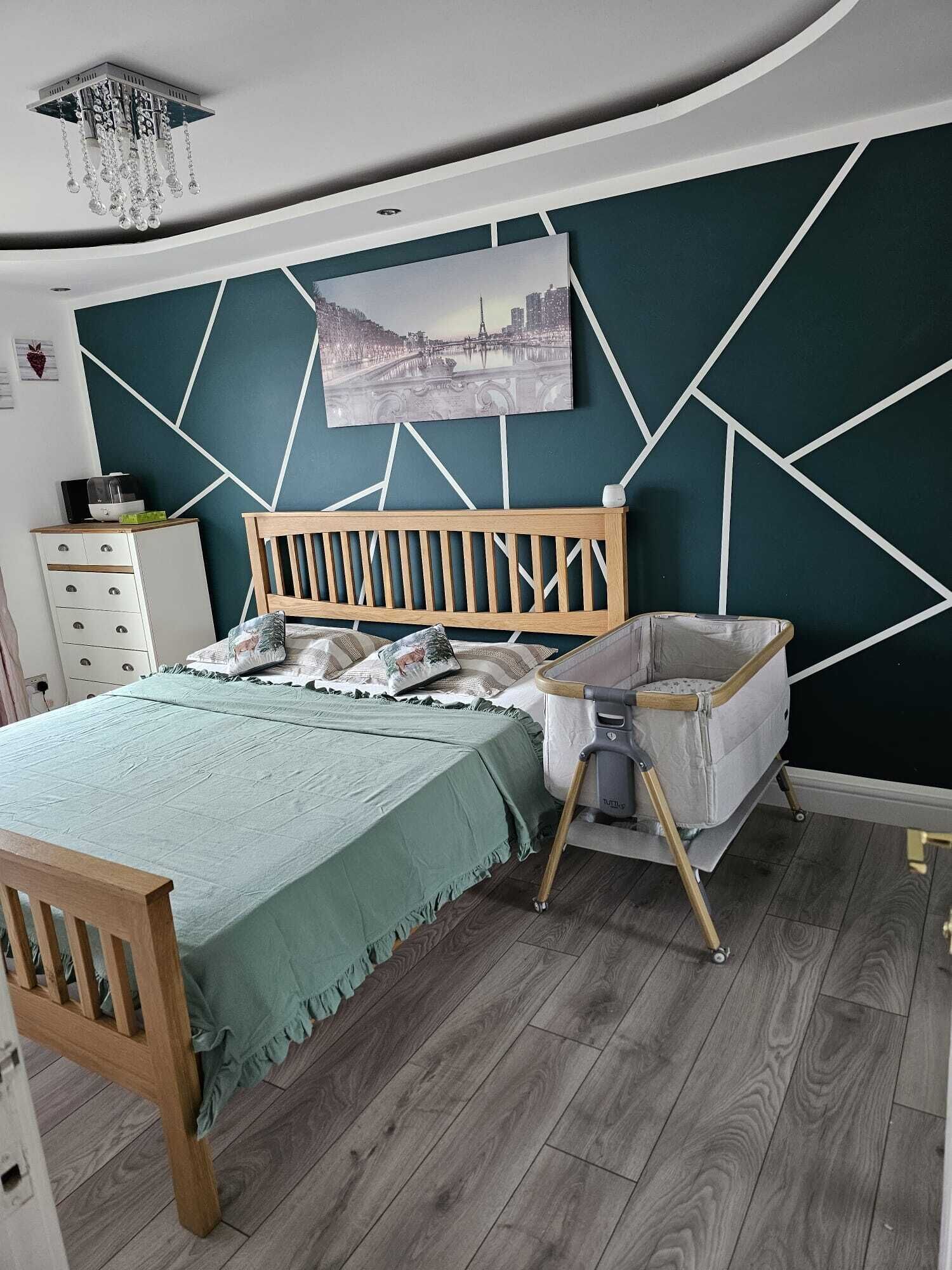 Ein Schlafzimmer mit einem Bett, einem Holzkopfstück, einem Babybett, einer Kommode und einer Wand mit geometrischen Mustern.