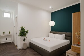 A modern bedroom with a large bed, a bedside table, a nightstand, a potted plant, and a wall-mounted lamp. - Grand Villa (Ho Chi Minh City)
