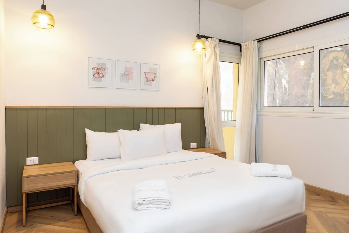 Un letto rifatto con cura, un asciugamano piegato e una coperta con il marchio, un comodino con una lampada e una finestra con tende.
