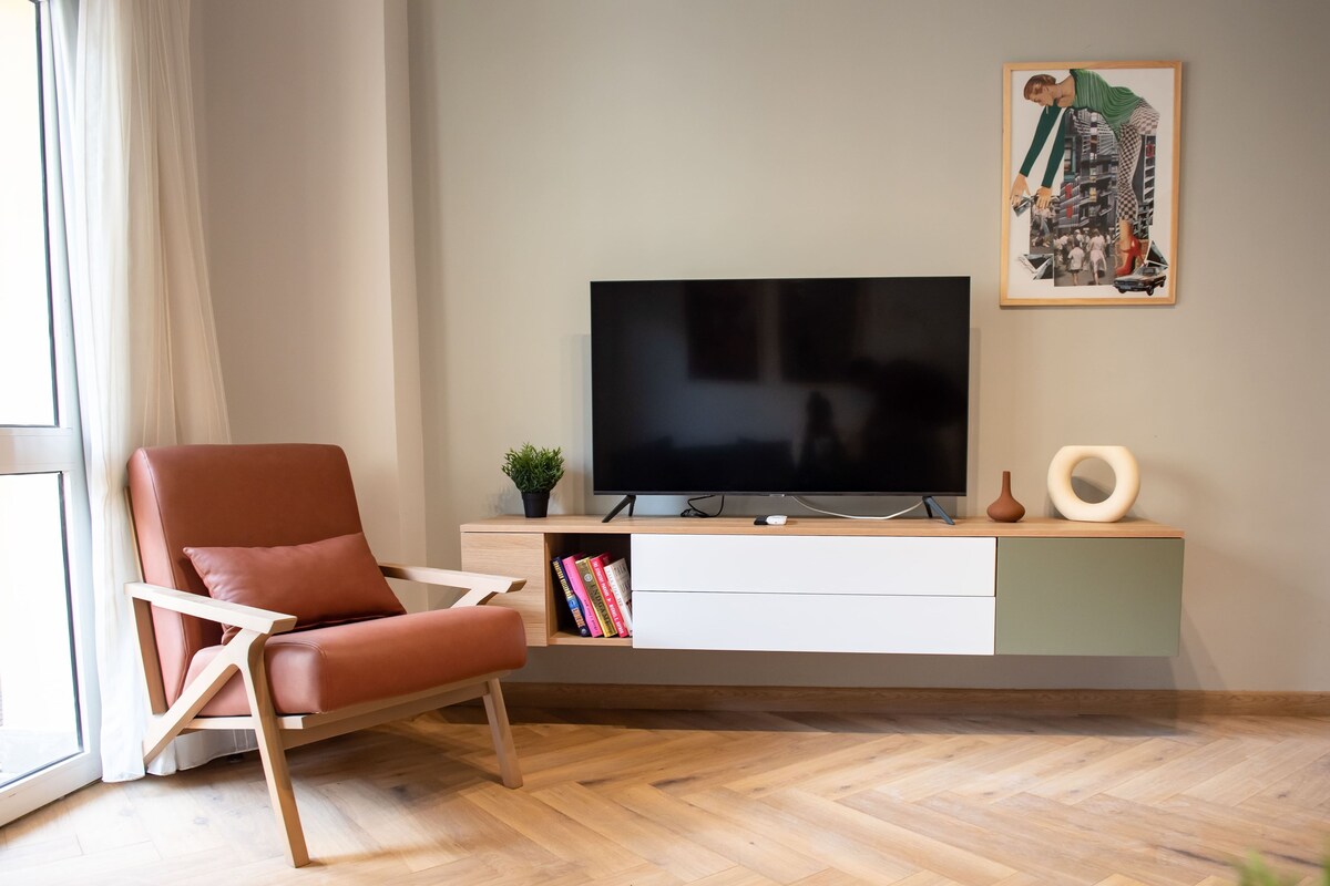 A modern living room with a flat-screen TV, a red leather chair, a wooden herringbone floor, and a minimalist console table with decorative items.