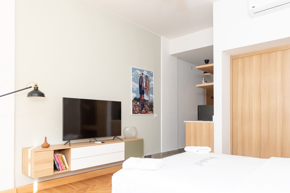 A modern hotel room with a flat-screen TV, a painting, a wooden wardrobe, and a bedside table.