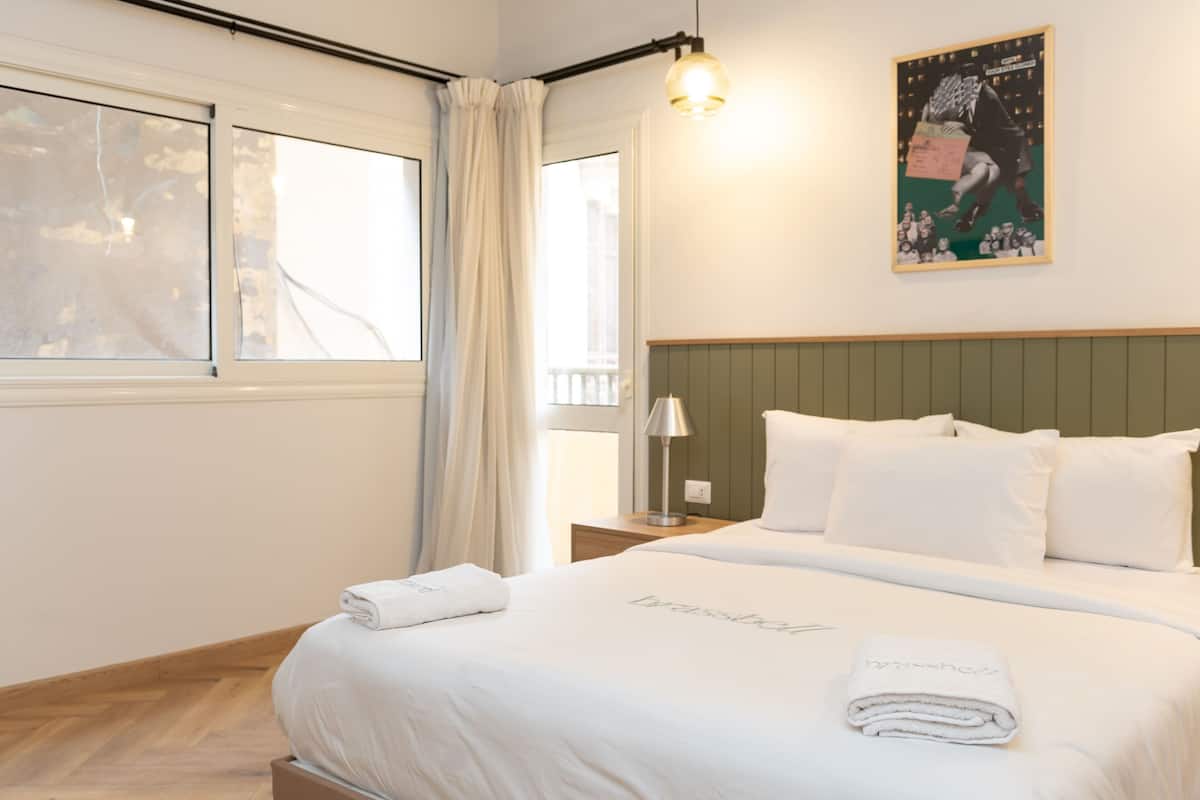 A hotel room with a bed, a nightstand, a lamp, a window with curtains, and a poster on the wall.
