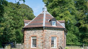 A stone building with a conical roof, two windows, and a chimney.