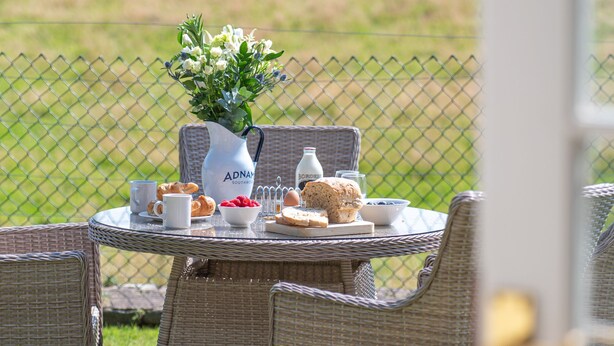 A wicker table set outdoors with a pitcher, cups, and pastries.