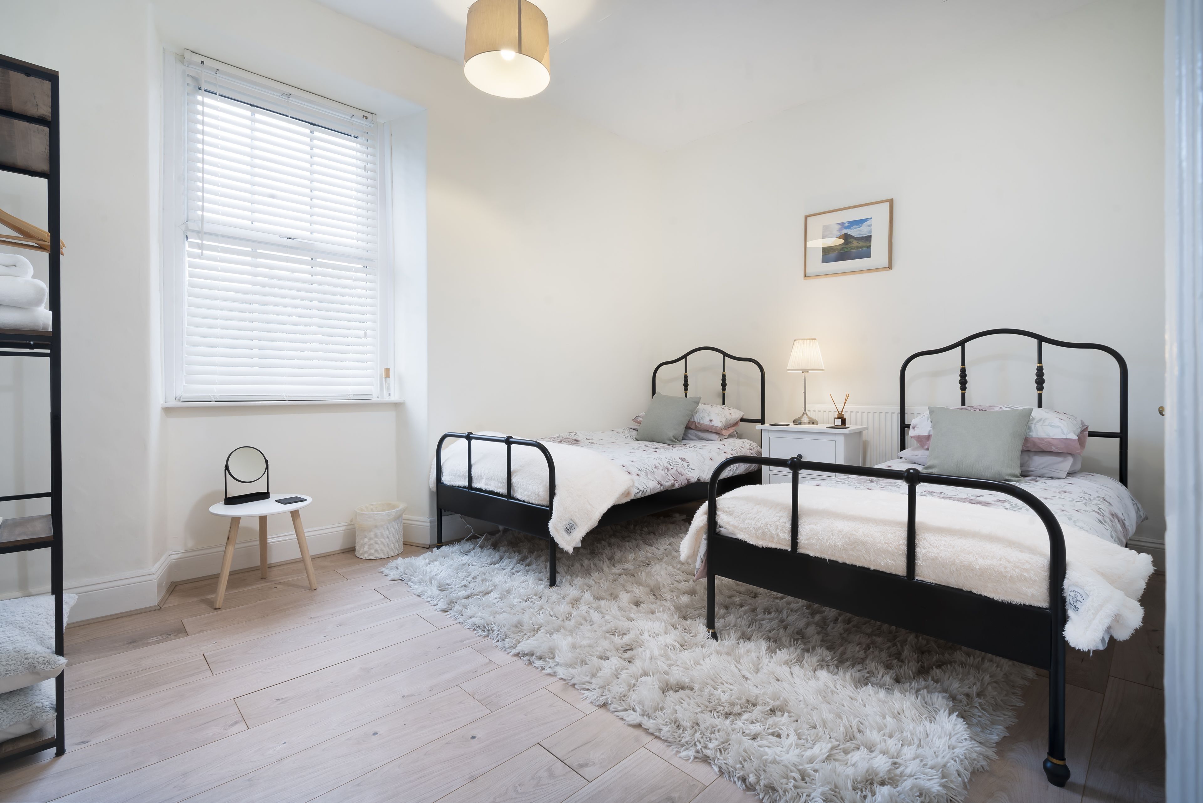 A bedroom with two black metal beds, a white fluffy rug, a small white chair, a bedside table with a lamp, and a framed picture on the wall.