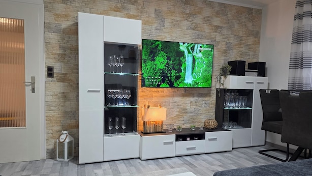 Ein modernes Wohnzimmer mit einer Steinwand, einem Flachbildfernseher, einer Glasvitrine mit Weingläsern und einem grauen Sofa.