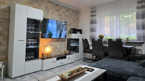 Ein modernes Wohnzimmer mit Steinwand, einem Flachbildfernseher, der über einem Glaskabinett angebracht ist, und einem Essbereich mit Tisch und Stühlen.
