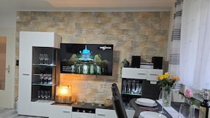 Ein modernes Wohnzimmer mit Steinwand, einem flachbildfernseher über einem Glastisch und einem Essbereich mit Tisch für zwei Personen.