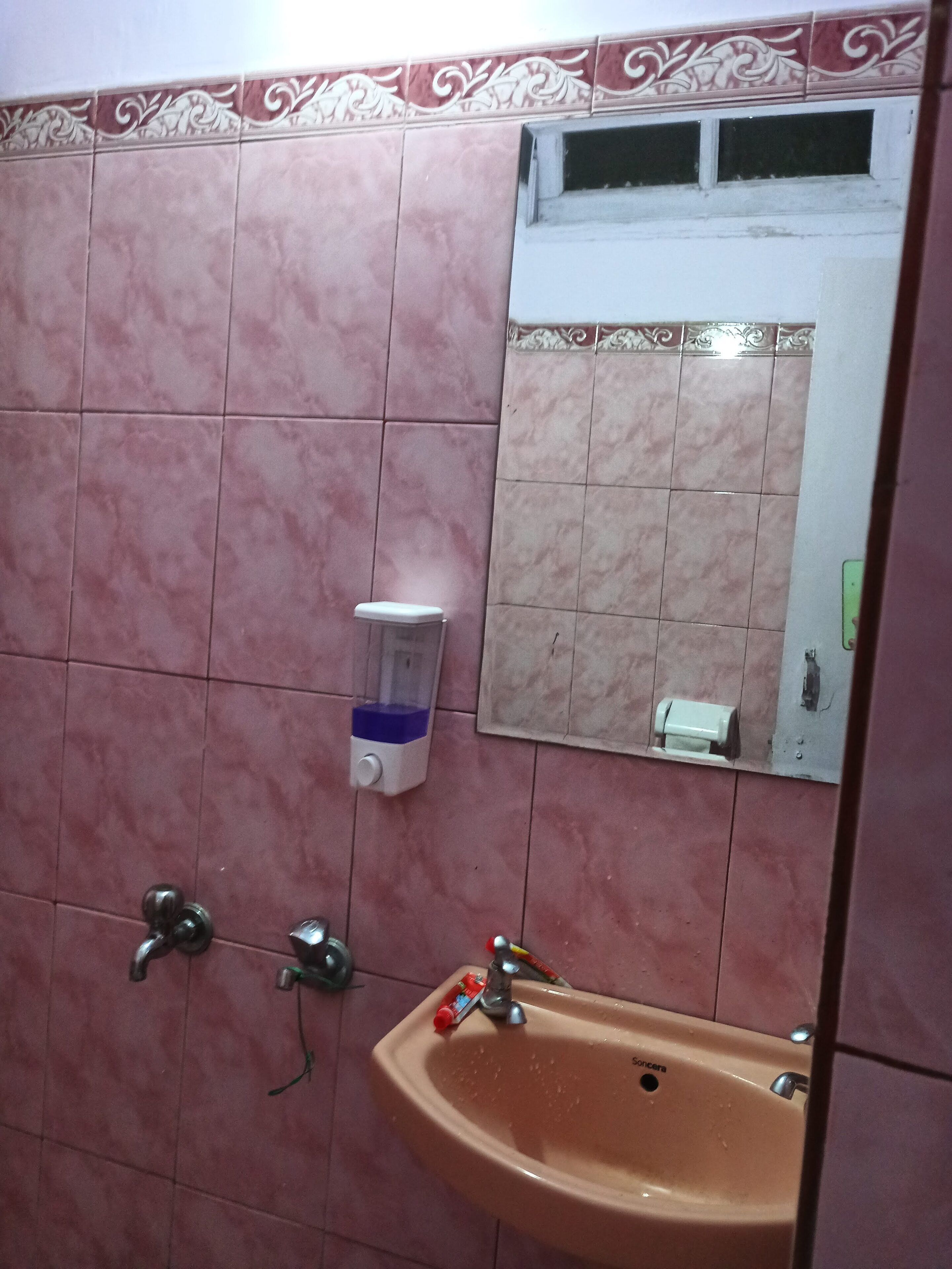 Un baño con azulejos rosas, un lavamanos, un espejo y un dispensador de jabón.