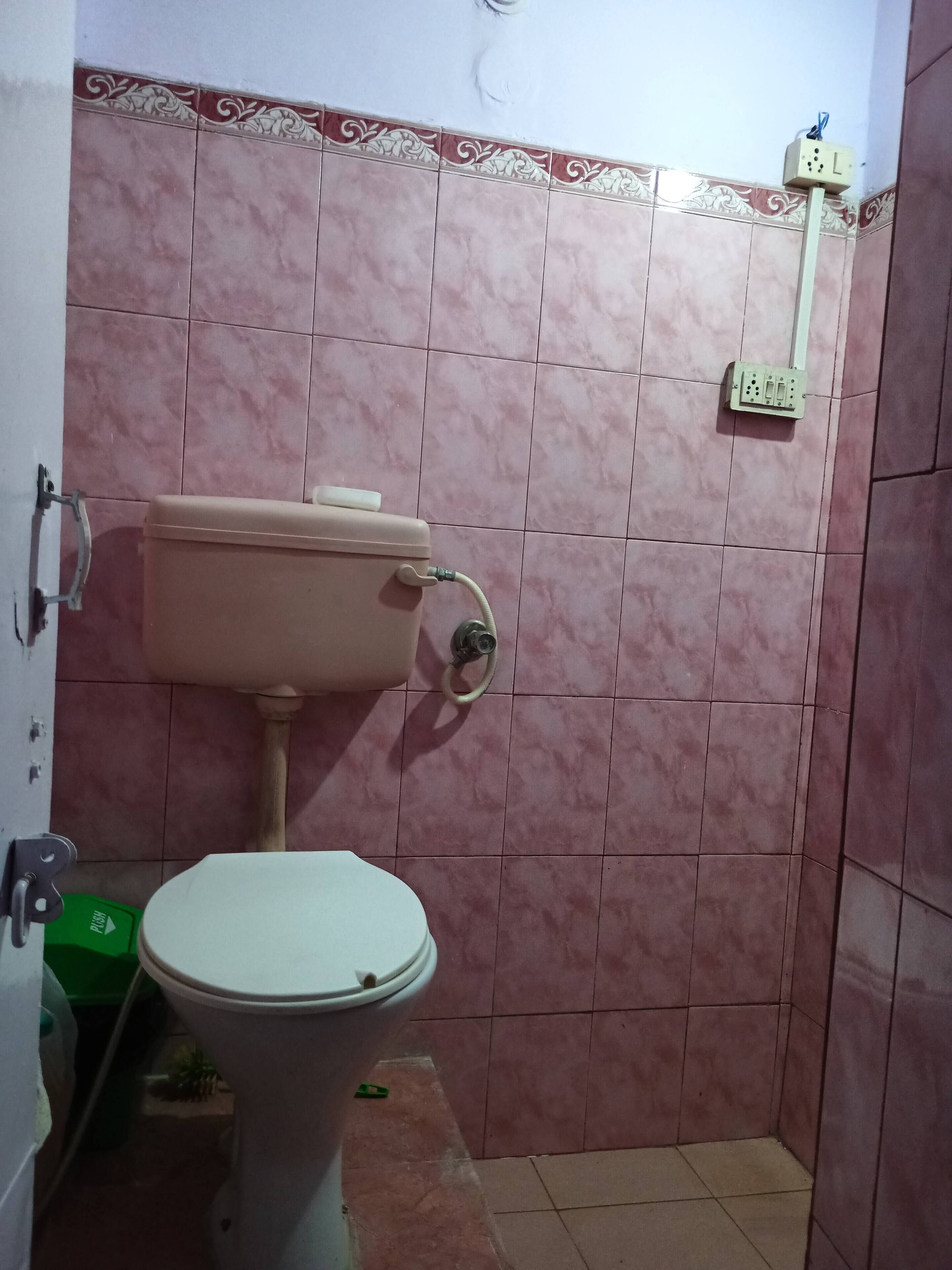 A small bathroom with pink tiles, a toilet, and a wall-mounted electrical outlet.
