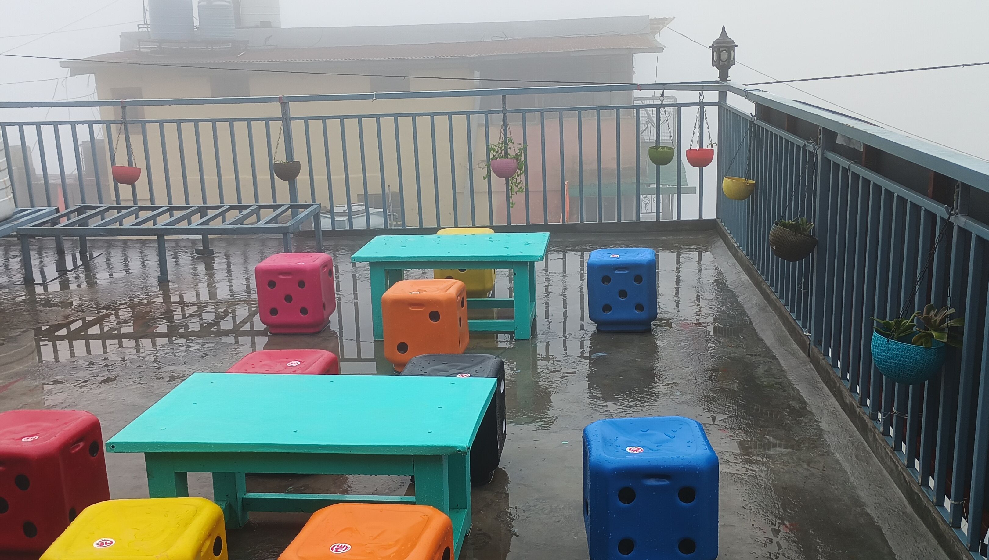 A wet balcony with colorful plastic stools and tables, a metal railing, and hanging planters.